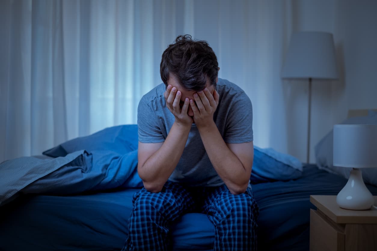 A man sitting on a bed with his face in his hands needs insomnia relief